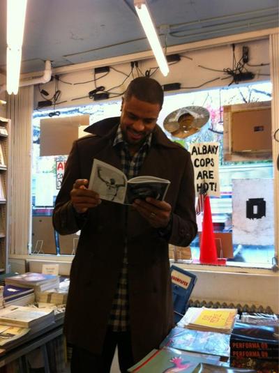 A person in a dark coat and checkered shirt stands in a cluttered, retro-style store, scanning an open book with a drawing on one page and text on the other. Behind them, a window displays signs including "ALBANY COPS ARE HOT!", alongside stacks of books on counters and a hanging police hat. Exposed wiring and flickering fluorescent lights add to the gritty atmosphere.