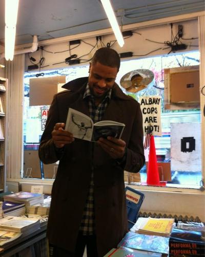 A person in a dark coat and checkered shirt stands in a cluttered, retro-style store, scanning an open book with a drawing on one page and text on the other. Behind them, a window displays signs including "ALBANY COPS ARE HOT!", alongside stacks of books on counters and a hanging police hat. Exposed wiring and flickering fluorescent lights add to the gritty atmosphere.