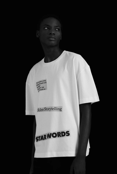 Person wearing white t-shirt with text patches: "Tmo artist, facing himsef w nst hs bld. To bld on.", "Storytelling", and bold black "STAR WORLDS" on chest, against flat black backdrop.
