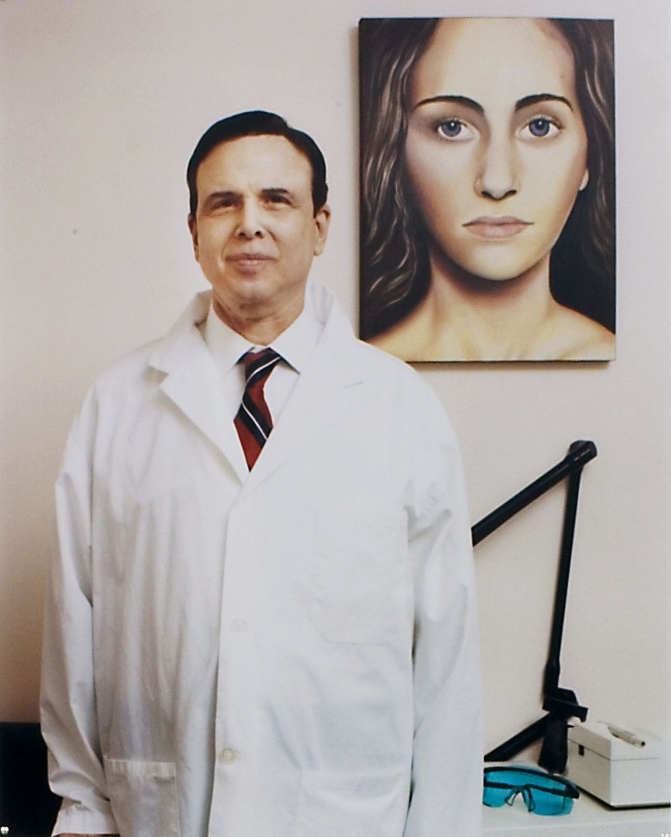 A man in a white lab coat, button-down shirt with a red and black striped tie, and subtle grin stands beside a large realistic painting of a nude woman with flowing dark hair and piercing blue eyes, in a clinical or medical office environment with neutral tones. His buttoned coat features functional pockets, and the painting contrasts formal and provocative elements.
