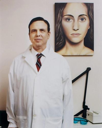 A man in a white lab coat, button-down shirt with a red and black striped tie, and subtle grin stands beside a large realistic painting of a nude woman with flowing dark hair and piercing blue eyes, in a clinical or medical office environment with neutral tones. His buttoned coat features functional pockets, and the painting contrasts formal and provocative elements.