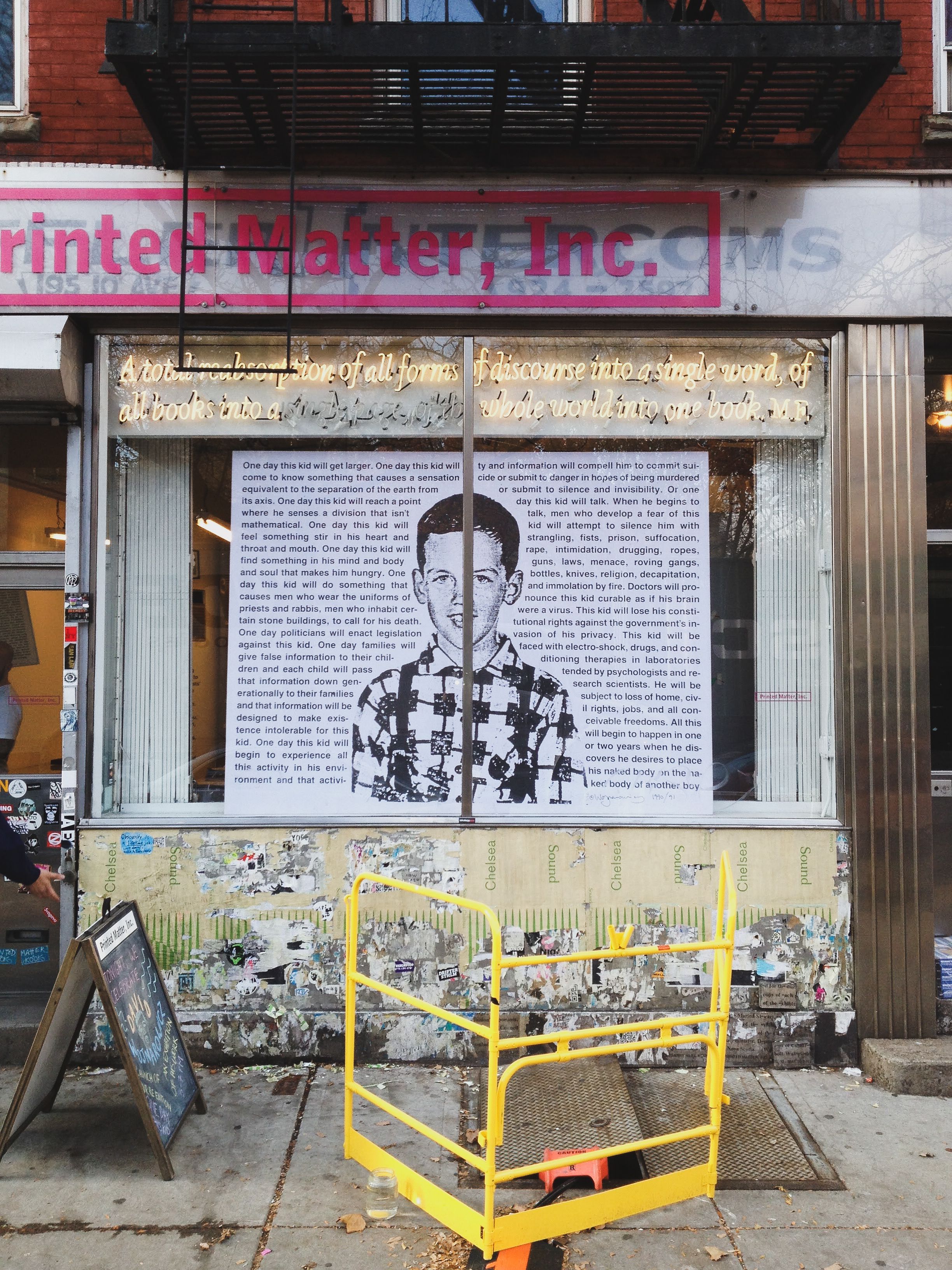 Printed Matter, Inc. storefront with neon signage featuring a quote about censorship and control by MF, large poster window displaying a boy’s portrait and underground texts, urban street scene with yellow barriers and construction signage.