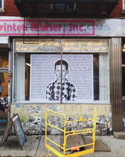 Printed Matter, Inc. storefront with neon signage featuring a quote about censorship and control by MF, large poster window displaying a boy’s portrait and underground texts, urban street scene with yellow barriers and construction signage.