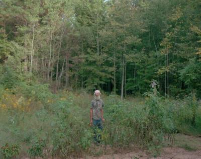 An older man with a white beard and patterned shirt stands among dense vegetation in a forest clearing, with tall trees in the background.