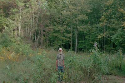 An older man with a white beard and patterned shirt stands among dense vegetation in a forest clearing, with tall trees in the background.