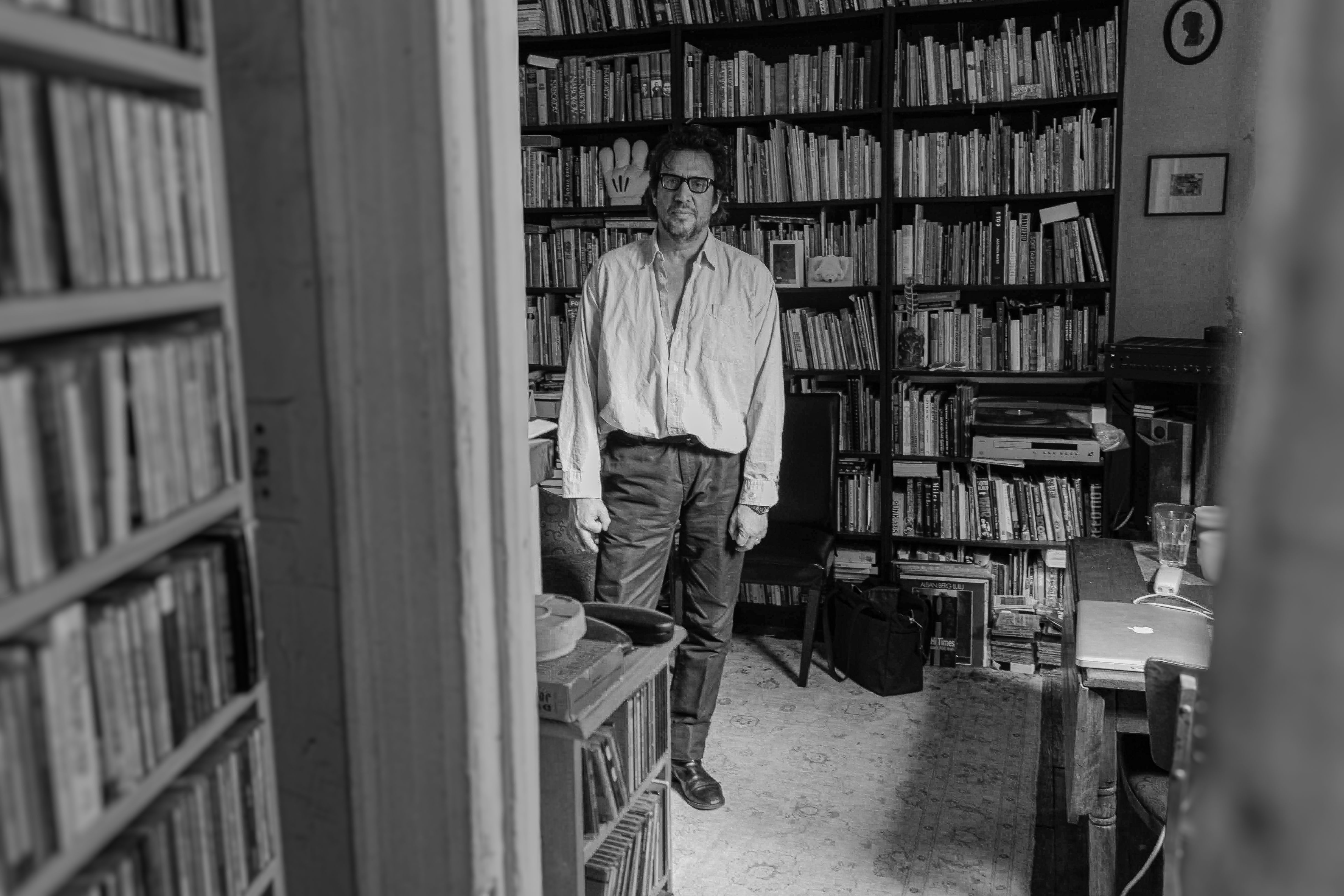A person stands in a cozy room filled with bookshelves, vintage electronics, and a patterned rug, smiling warmly at the camera.