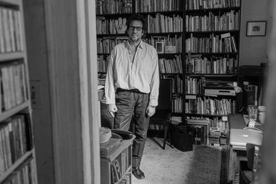 A person stands in a cozy room filled with bookshelves, vintage electronics, and a patterned rug, smiling warmly at the camera.