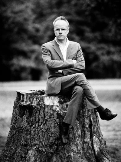 A man in glasses, a suit, and tie sits confidently on a tree stump with crossed arms, outdoors against a blurred greenery backdrop.