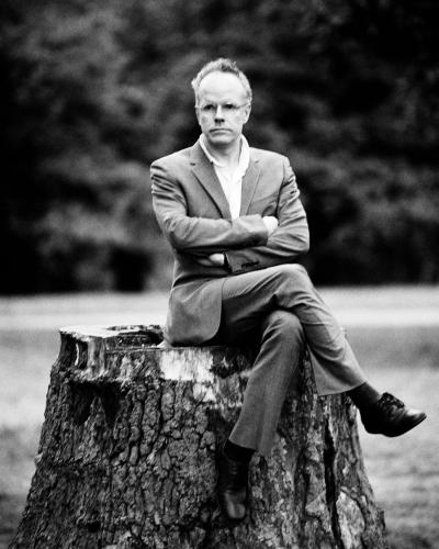 A man in glasses, a suit, and tie sits confidently on a tree stump with crossed arms, outdoors against a blurred greenery backdrop.