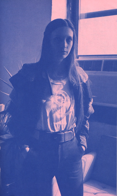 Woman with long hair, wearing a jacket and t-shirt with a logo, stands with hands in pockets.