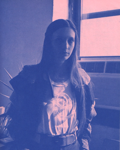 Woman with long hair, wearing a jacket and t-shirt with a logo, stands with hands in pockets.