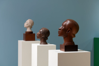 Three sculpted busts arranged on cream pedestals against a solid blue wall. Foremost: a tan marble head on a dark wooden base, slender form. Middle: a dark bronze bust with textured surface and elongated neck. Rightmost: a glossier bronze figure with rounded contours, minimal detailing. Green panel visible on far right.