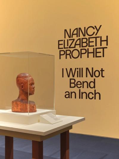 Wooden sculpture in a glass display case mounted on a white pedestal against a mustard-yellow wall with "I Will Not Bend an Inch" in dark bold text, atop a small informational stand, with brown pedestal legs against a slate-gray floor.