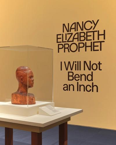 Wooden sculpture in a glass display case mounted on a white pedestal against a mustard-yellow wall with "I Will Not Bend an Inch" in dark bold text, atop a small informational stand, with brown pedestal legs against a slate-gray floor.