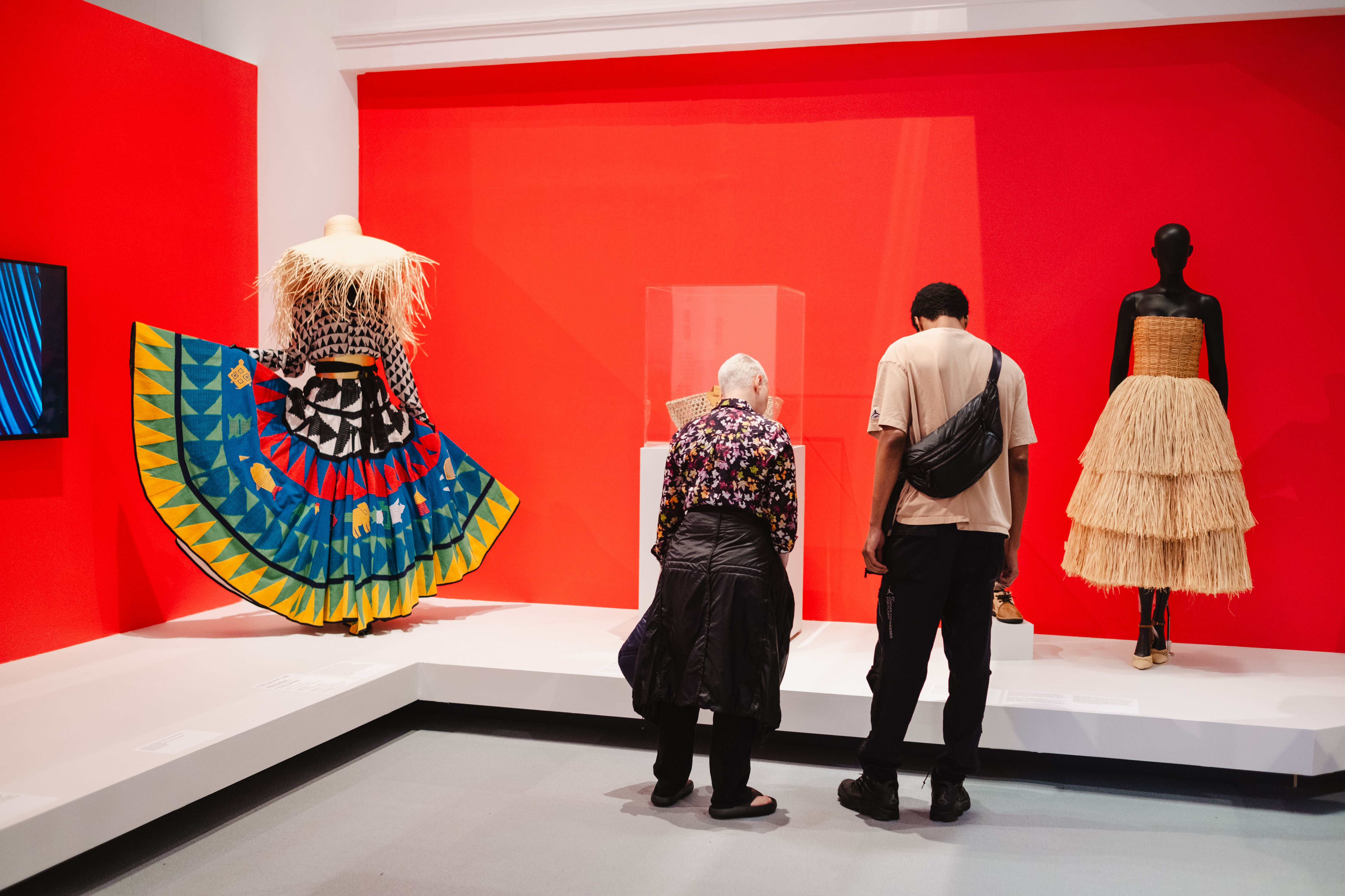 Two people view colorful, layered dresses on mannequins against a bright red wall.