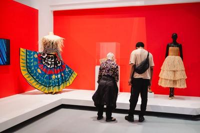 Two people view colorful, layered dresses on mannequins against a bright red wall.