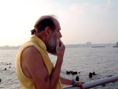 Gray-bearded man wearing yellow tunic on pier overlooking calm water and hazy city skyline at sunset.