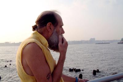Gray-bearded man wearing yellow tunic on pier overlooking calm water and hazy city skyline at sunset.