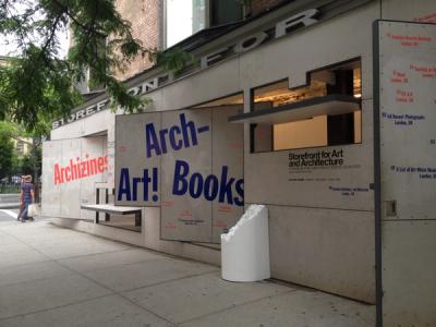 The image depicts a storefront at 97 Kenmare Street, New York, NY, with concrete panels attached to its exterior, advertising various art and architecture programs and exhibitions. Prominent text includes "Archizines" in red, "Arch-Art! Books" in purple, and "Storefront for Art and Architecture" in black along with gallery hours and address details. A white cylindrical sculpture sits near the entrance. A brick building, tree branches, and a person with a bag and shawl are visible nearby.