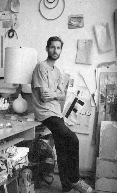 Man with beard, arms crossed, sits on desk in cluttered art studio with fan and artwork.