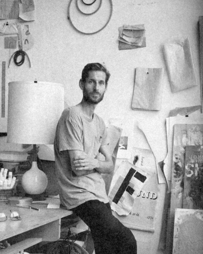 Man with beard, arms crossed, sits on desk in cluttered art studio with fan and artwork.