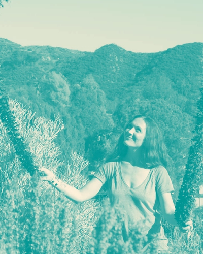A person forms a circular wreath with arms, standing against soft green-toned hills and trees in an ethereal outdoor setting, evoking a dreamy, organic atmosphere through warm lighting and vintage filters.