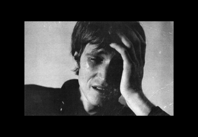 Person with short dark hair covers face with left hand, eyes partially closed, against plain light wall under soft lighting. Somber black and white photograph.
