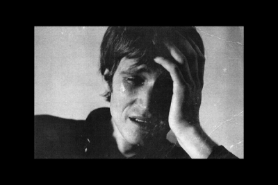 Person with short dark hair covers face with left hand, eyes partially closed, against plain light wall under soft lighting. Somber black and white photograph.