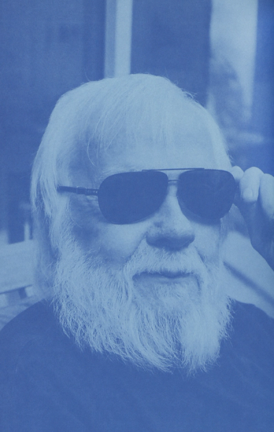 Elderly man with long white beard and hair, wearing aviator sunglasses, adjusts them with his hand.