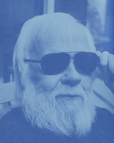 Elderly man with long white beard and hair, wearing aviator sunglasses, adjusts them with his hand.