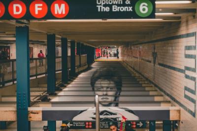 NYC subway station with BDFM and 6 signs, tiled walls, and a large poster of a face on stairs.