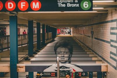 NYC subway station with BDFM and 6 signs, tiled walls, and a large poster of a face on stairs.