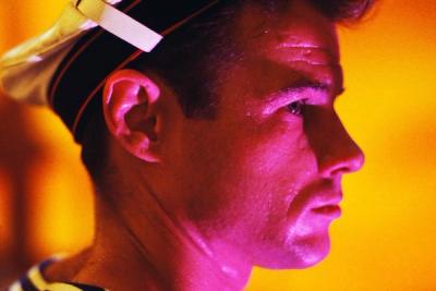 A young man in profile, wearing a white uniform cap with a black band and red interior, illuminated by warm orange tones, ear and eyes visible, blurred orange background.