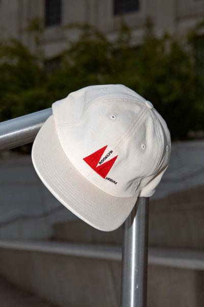 White baseball cap with red and black "Brooklyn Usetum" logo on front, resting on metal railing outdoors.