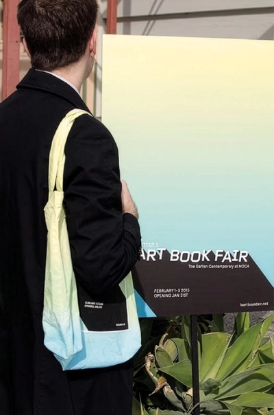 Man in black coat with ombre tote bag faces sign for "ART BOOK FAIR"