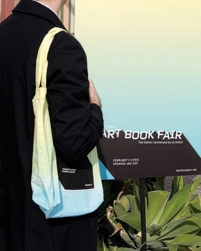 Man in black coat with ombre tote bag faces sign for "ART BOOK FAIR"