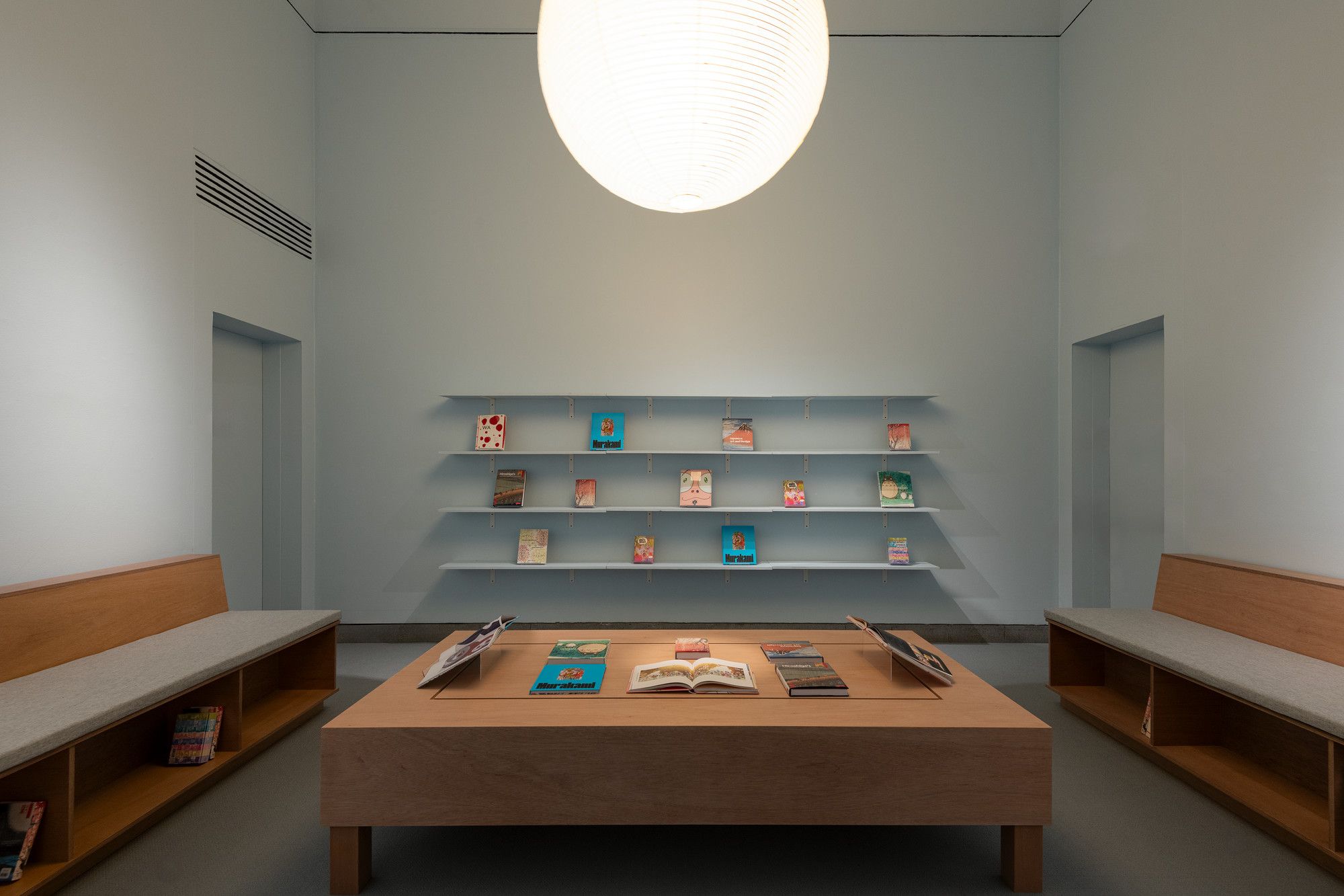 Minimalist room with books on shelves and tables, light blue walls, wooden benches, and a large pendant light.
