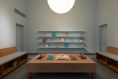Minimalist room with books on shelves and tables, light blue walls, wooden benches, and a large pendant light.