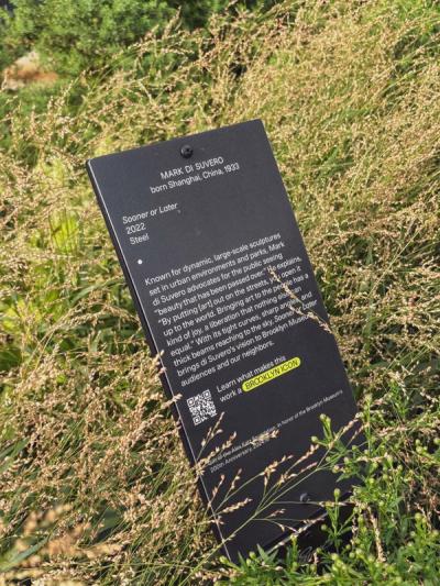 Plaque for Mark Di Suvero (1933–2022), Shanghai-born steel artist. White text on black: dynamic sculptures in parks/streets. QR code 'BROOKLYN ICON.' Brooklyn Museum's 200th Anniversary.