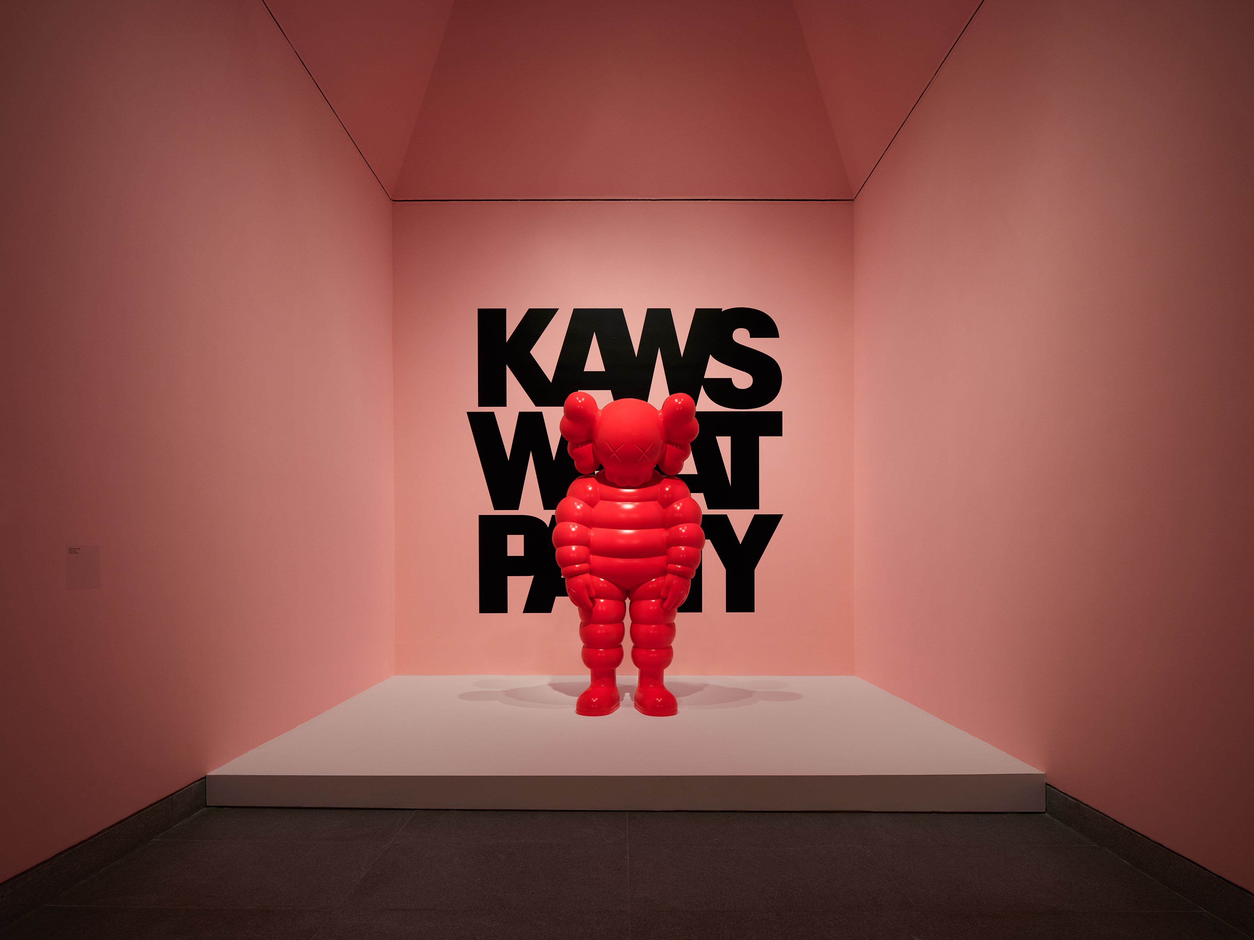 Red KAWS Companion figure stands before black text on a pink wall.