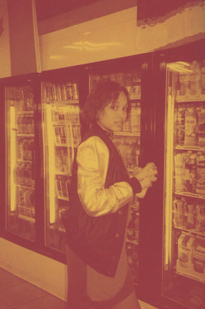Woman in bomber jacket holds can in front of refrigerated drink display.