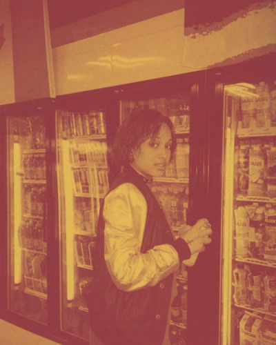 Woman in bomber jacket holds can in front of refrigerated drink display.