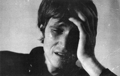 Person with short dark hair covers face with left hand, eyes partially closed, against plain light wall under soft lighting. Somber black and white photograph.