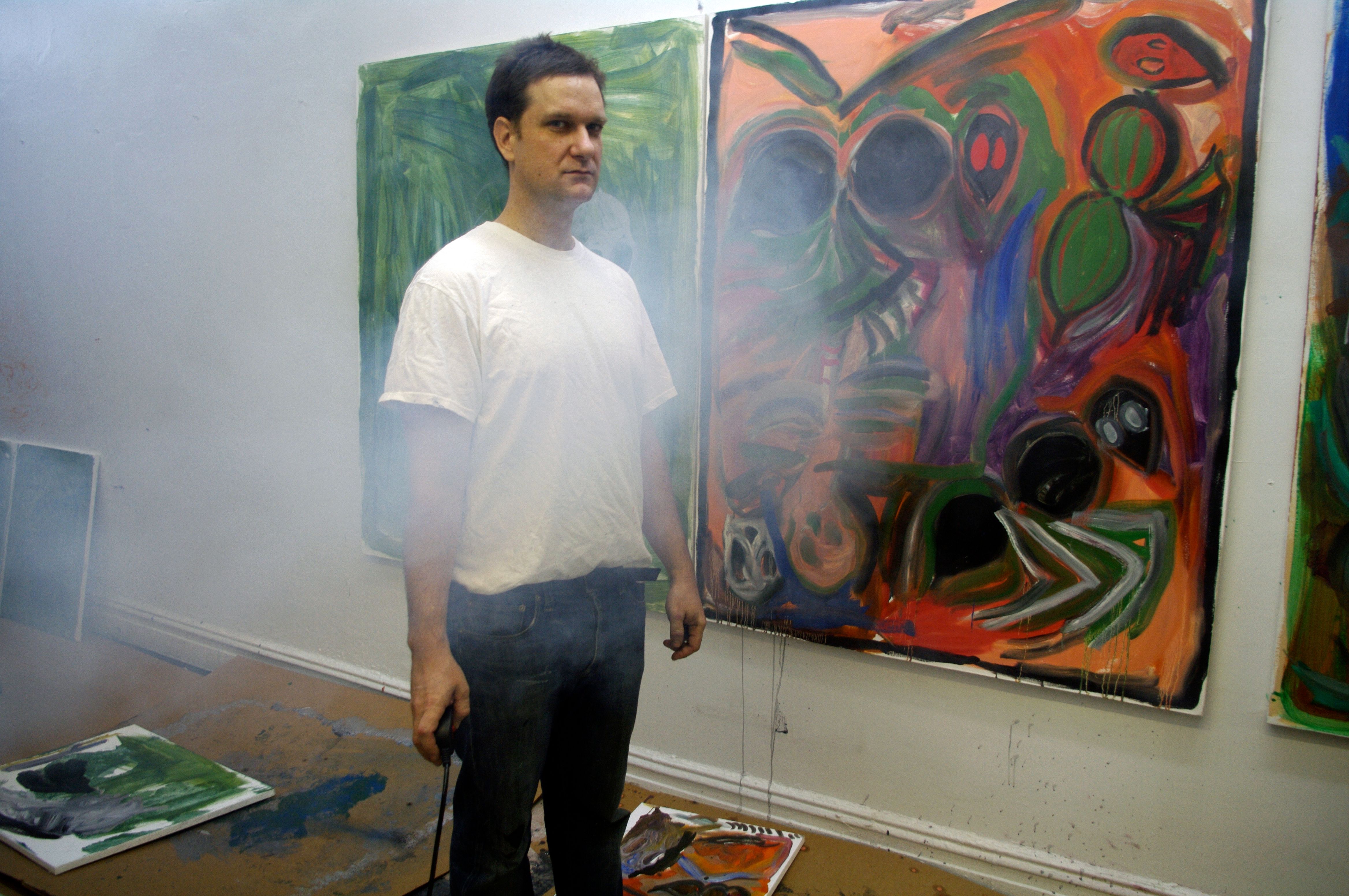 A man in a white t-shirt and blue jeans stands in an art space surrounded by abstract paintings with vibrant colors and splattered textures. Paintings lie on the unfinished plywood floor, including a large green canvas. The environment is hazy with visible smudges and paint splatters on the walls.