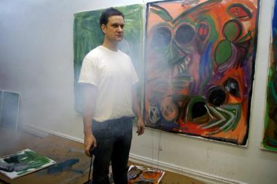 A man in a white t-shirt and blue jeans stands in an art space surrounded by abstract paintings with vibrant colors and splattered textures. Paintings lie on the unfinished plywood floor, including a large green canvas. The environment is hazy with visible smudges and paint splatters on the walls.