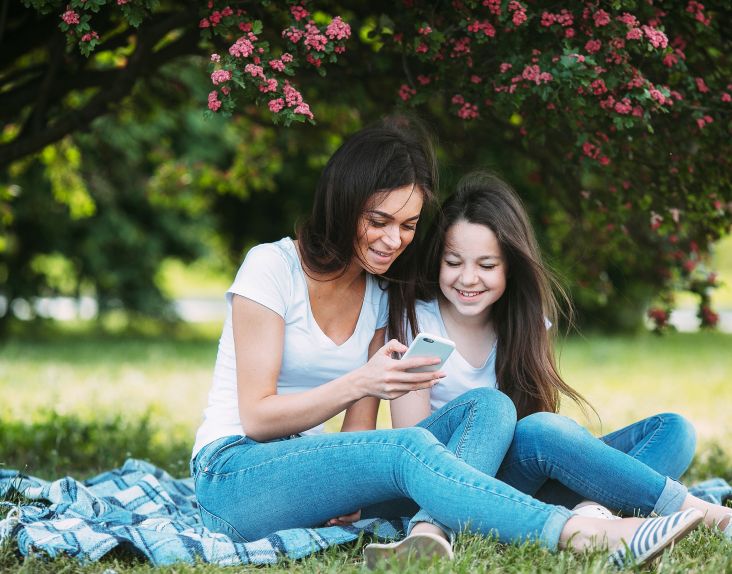 Mor og datter sitter på en gresslette under ett tre og ser på en mobiltelefon