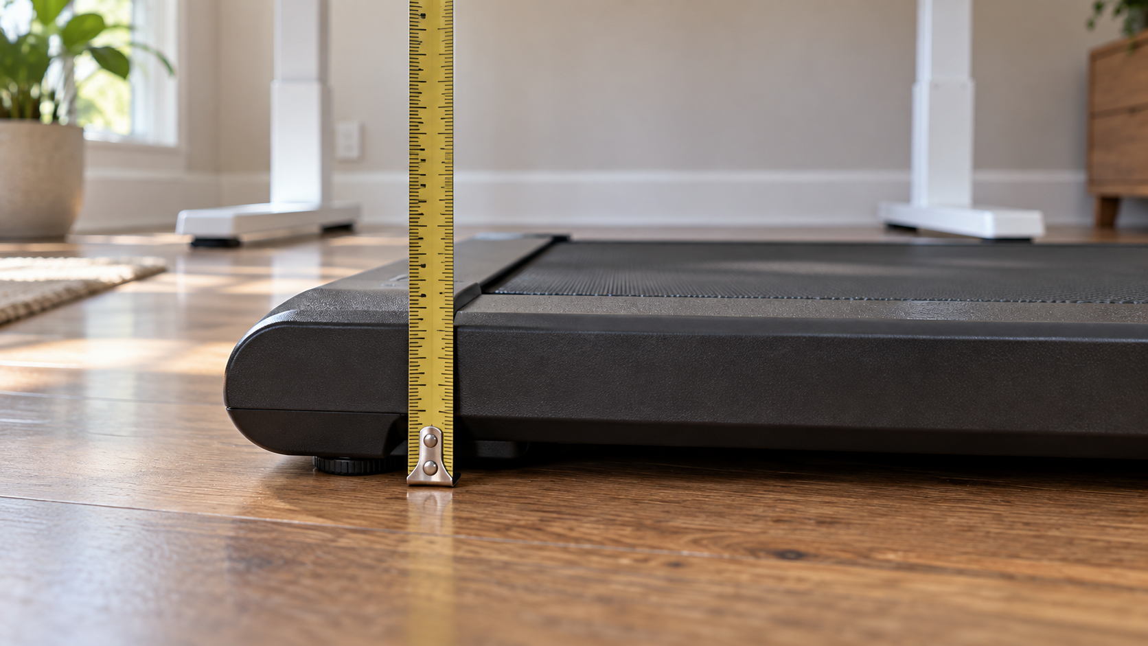 A tape measure held upright against the edge of a walking pad's deck on a wood floor, showing the deck's height, with the legs of a standing desk behind it.