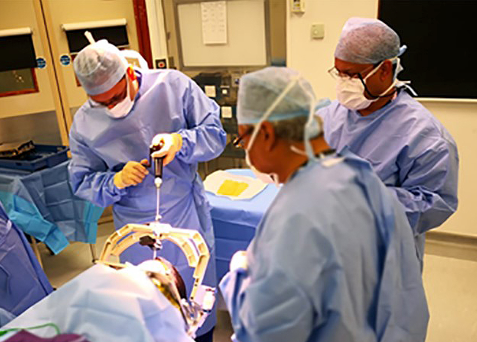 Dr Pereira drilling a hole in a patient's skull while Profs Nandi and Aziz look on