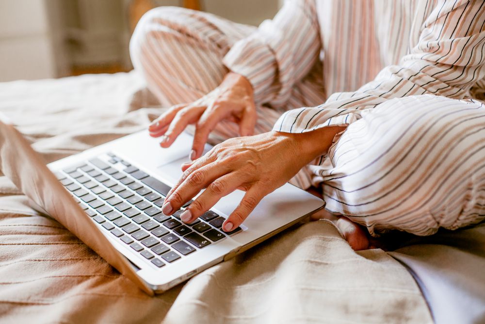 Elderly woman sitting on bed in pyjamas, typing on laptop