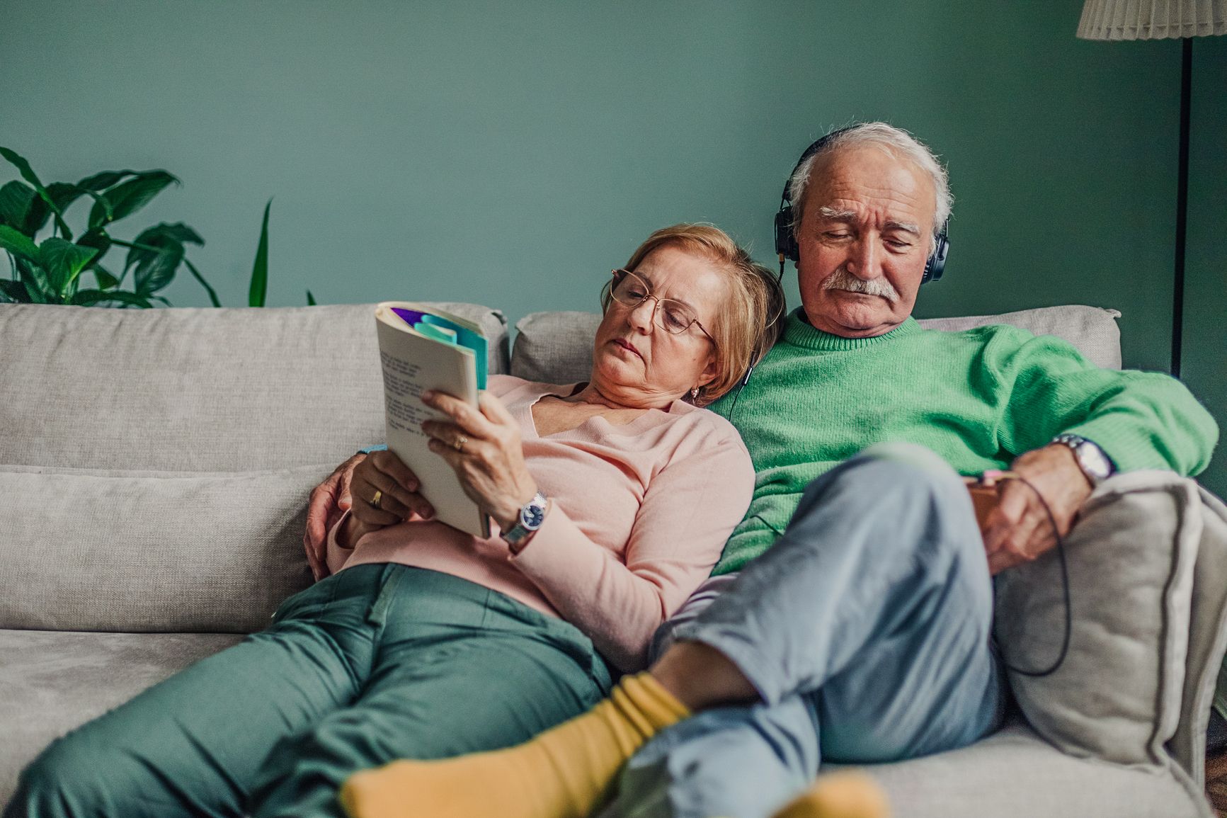 Older couple relaxing together on the couch. Woman is reading and comfortably leaning against the man who is listening to headphones with him arm around her..